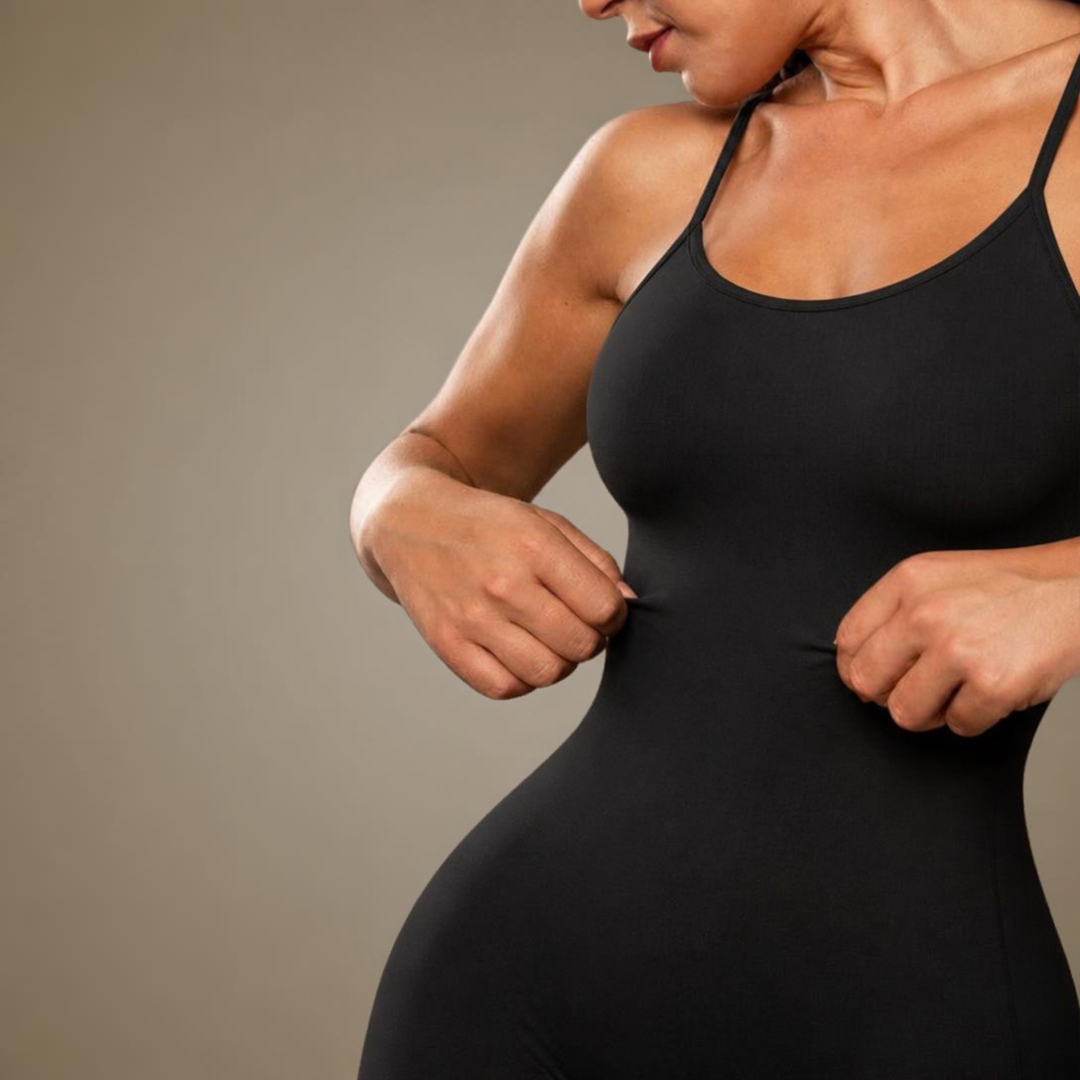 A close-up of a woman wearing a black full body tummy-control bodysuit shapewear by BodyFlexx pulling the fabric slightly at the waist against a plain taupe background.