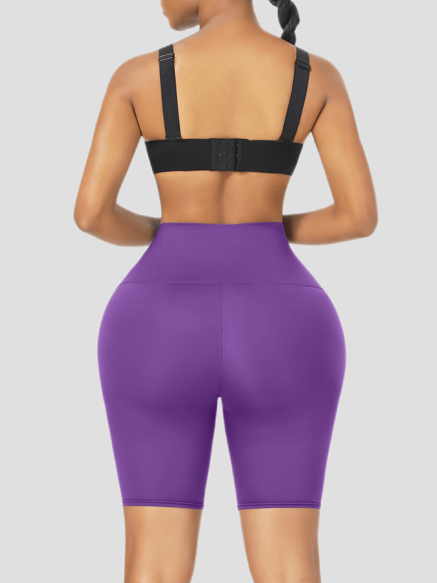 Woman standing backwards wearing high-waisted purple sauna activewear shorts and a black sports top smiling after her workout.