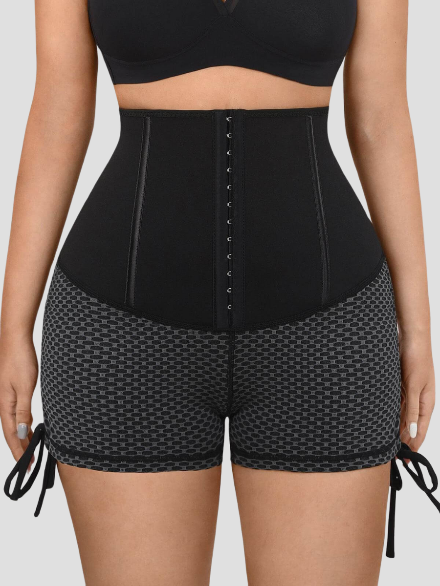 Close up front view of a woman wearing black and gray patterned sauna sweat belt lined activewear shorts with a corset-style waist training closure.