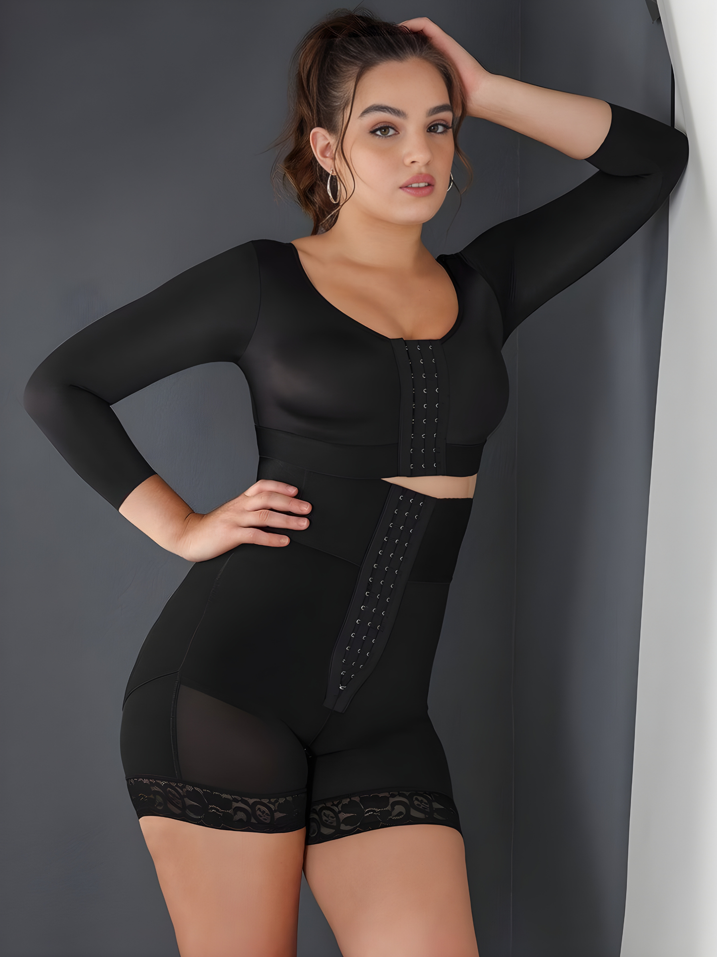 Woman wearing a black Columbian Faja waist-training corset shapewear garment by BodyFlexx leaning against a gray wall.