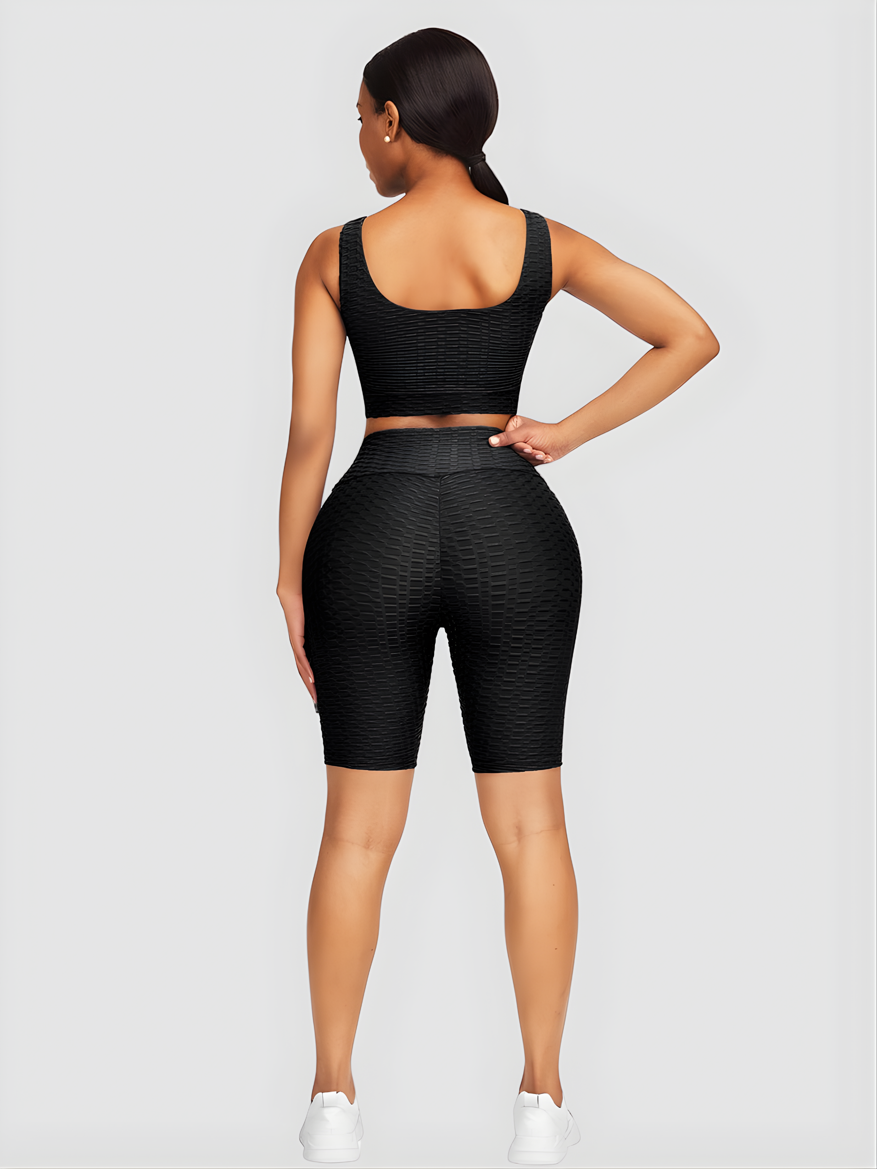 Woman wearing a black activewear set with a matching sports bra and high-waisted tummy control mid-thigh spandex shorts. She is standing backwards with her hand on her hip showing off the backside curves and scrunch bum design.