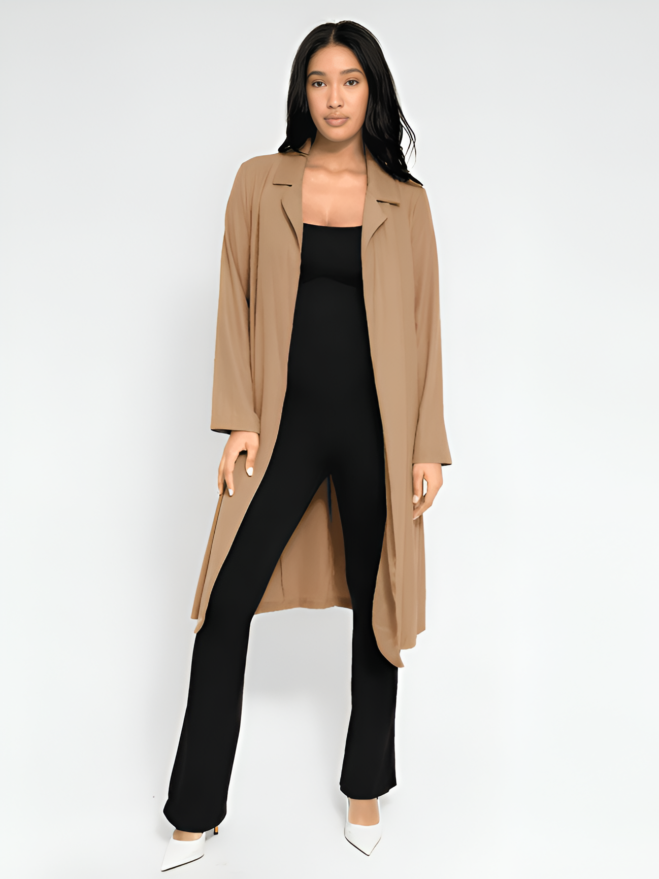 Woman wearing a beige trench coat over a black shapewear jumpsuit with flared legs and white heels.