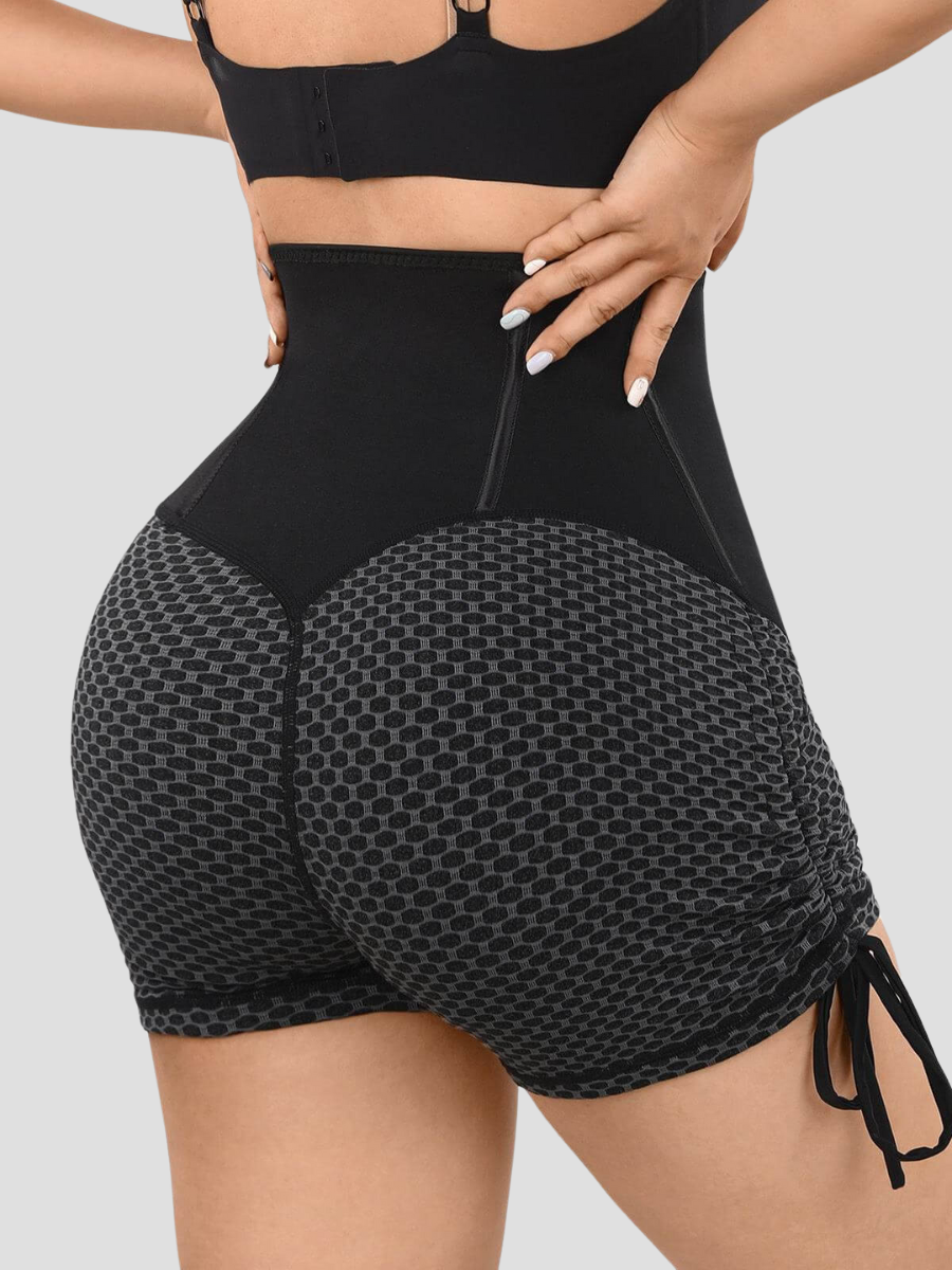 Woman standing backwards wearing sauna activewear shorts lined with neoprene with a black and gray honeycomb pattern and a corset-style hook-and-eye closure.